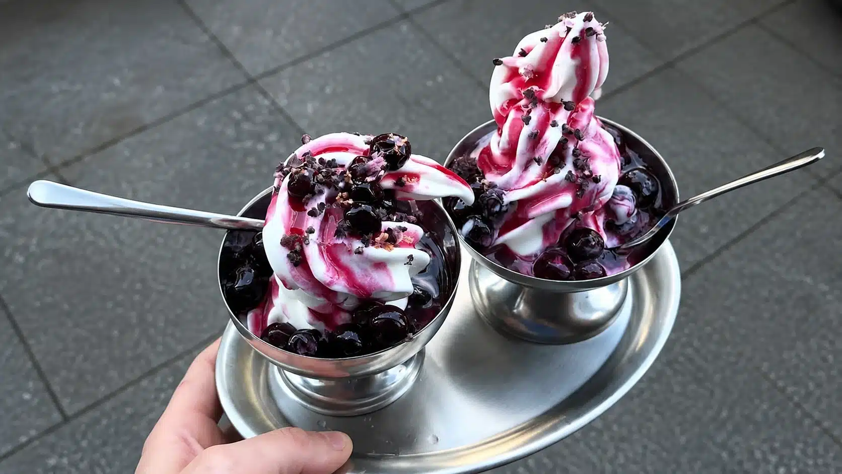 Two berry soft serve desserts in metal cups topped with whipped cream, berry sauce, and chocolate crumbs, served outdoors on a tray