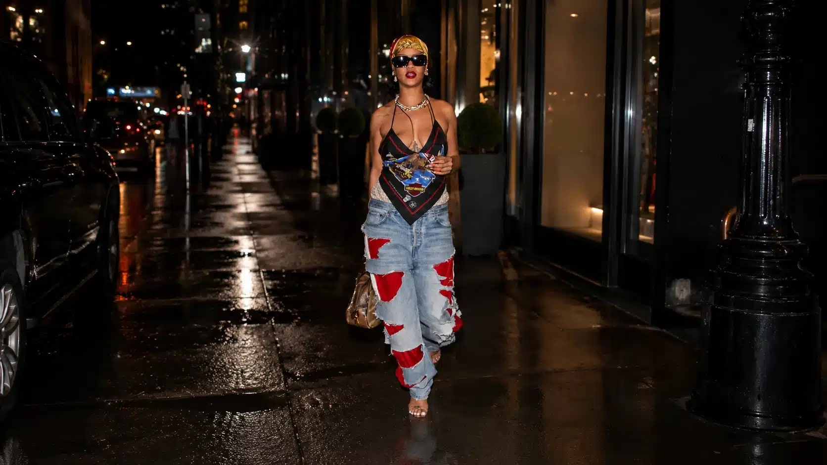 woman in scarf top and ripped jeans walks on wet nighttime city sidewalk with sunglasses and gold shoulder bag heels