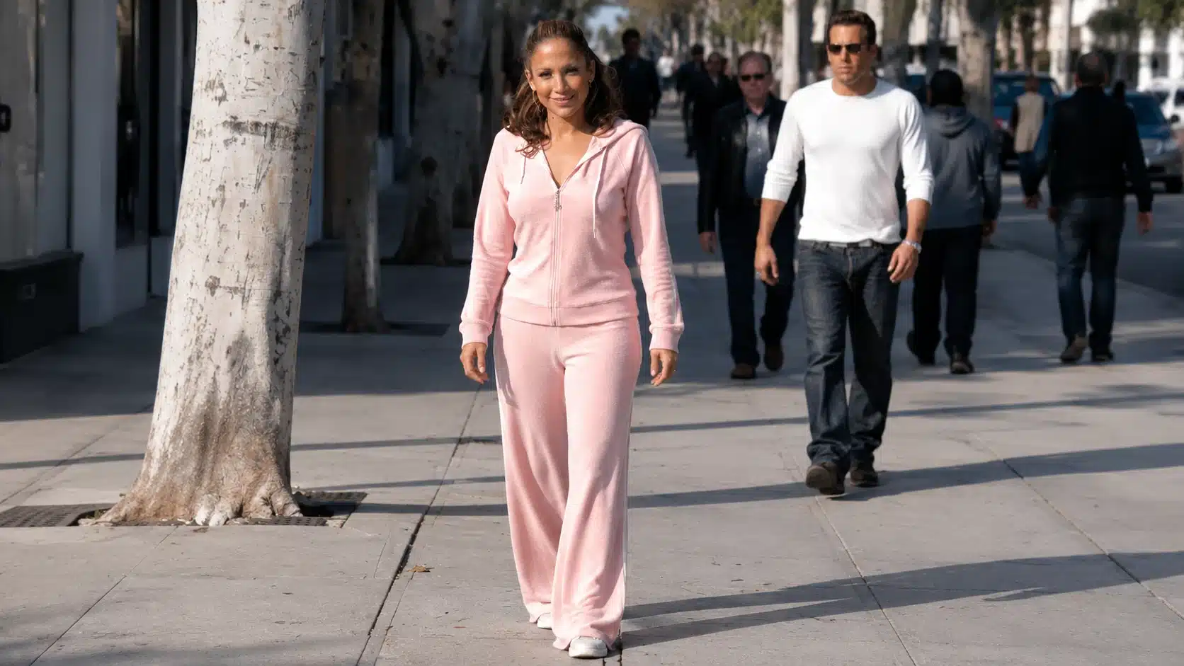 woman wearing pink velour tracksuit walks on sunny sidewalk with blurred pedestrians behind her in casual street style