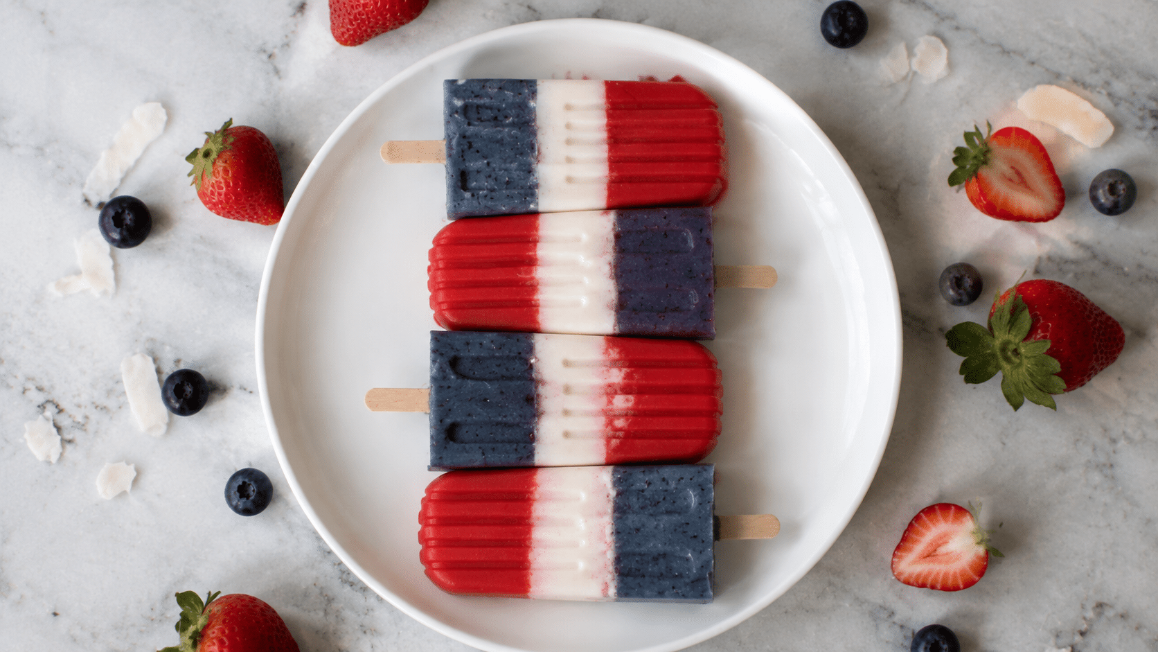 Four red, white, and blue popsicles arranged on a white plate, surrounded by fresh strawberries and blueberries on a light marble surface