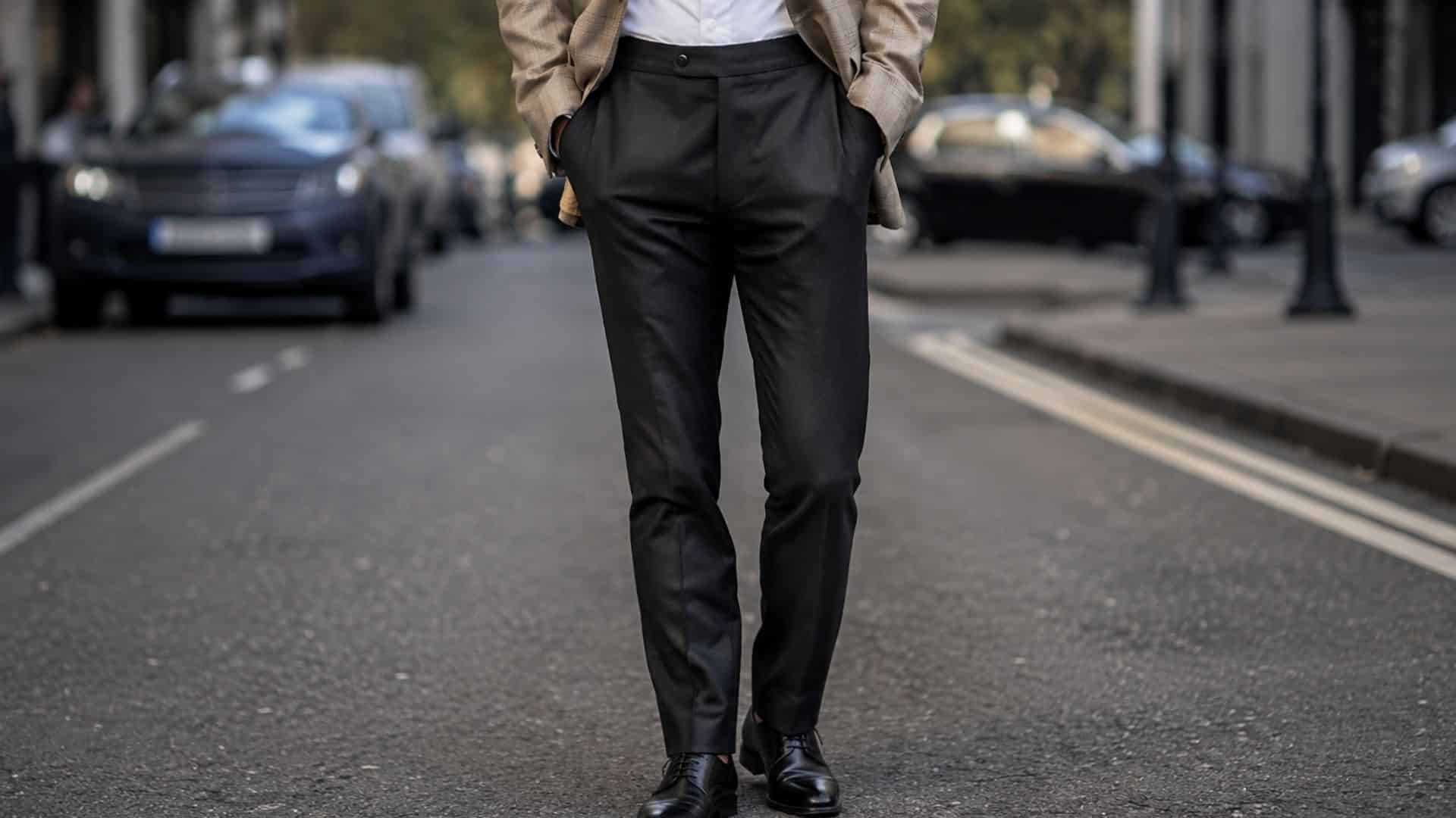 Man wearing tailored black trousers and a beige blazer while walking along a city street with parked cars in the background