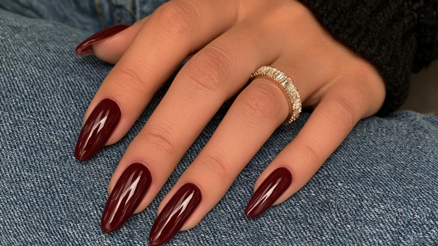 Hand with long, almond-shaped glossy deep burgundy-red manicured nails wearing a gold band ring.