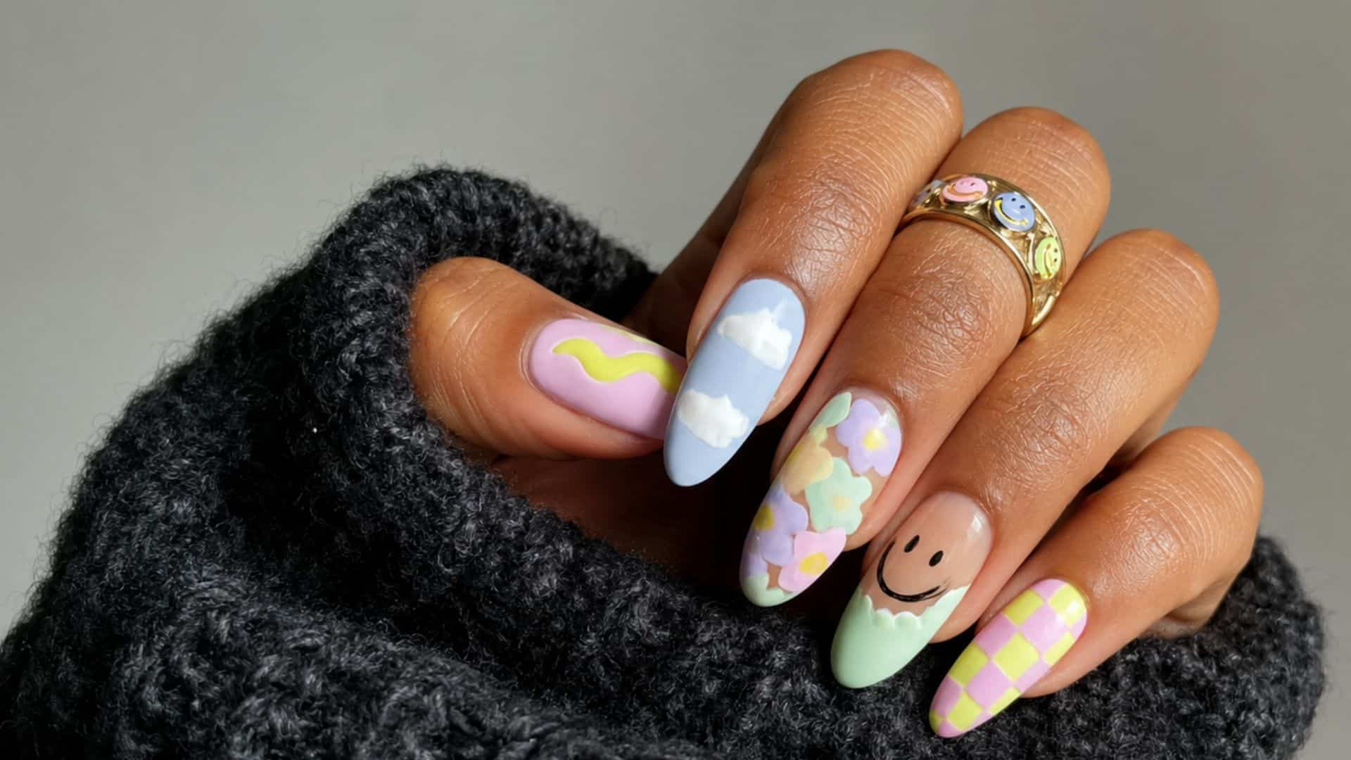 Pastel almond-shaped nails with cloud, floral, smiley face, checkerboard, and abstract swirl nail art on a hand.
