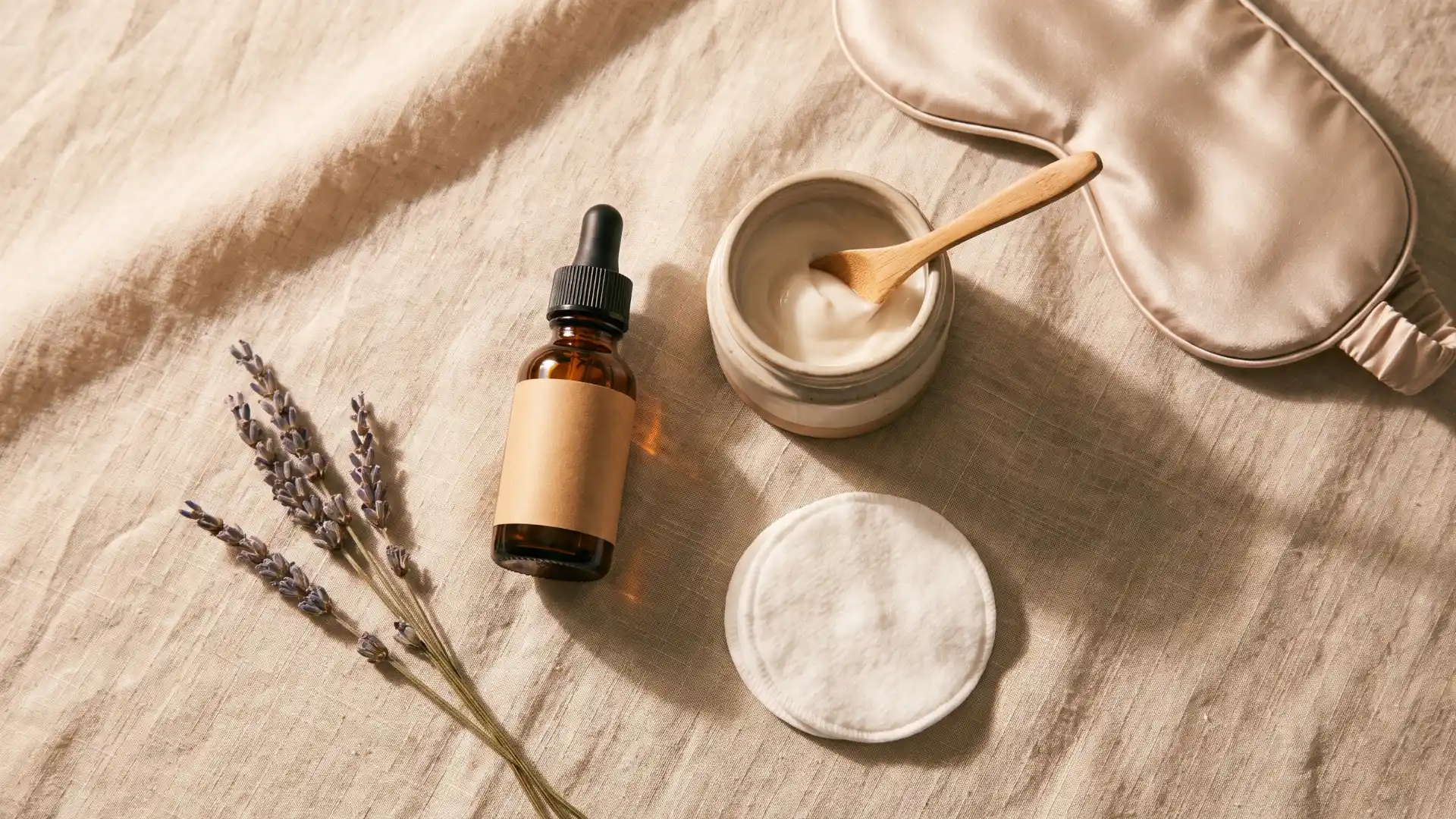 Skincare arrangement with serum bottle moisturizer cotton pad lavender and sleep mask on soft fabric background